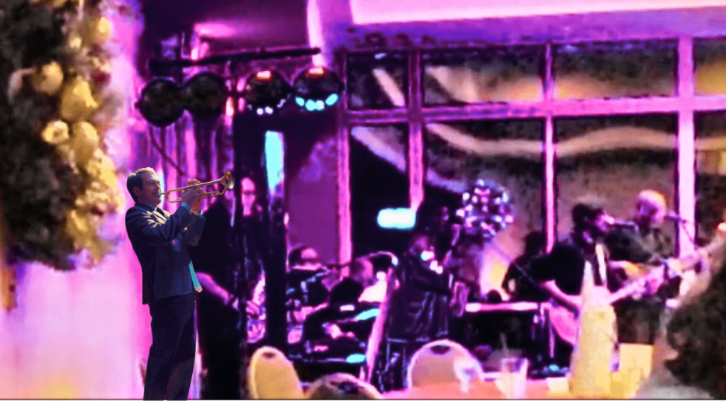 A live band performance featuring musicians playing various instruments, including a prominent trumpeter in a suit, with stage lights illuminating the group.