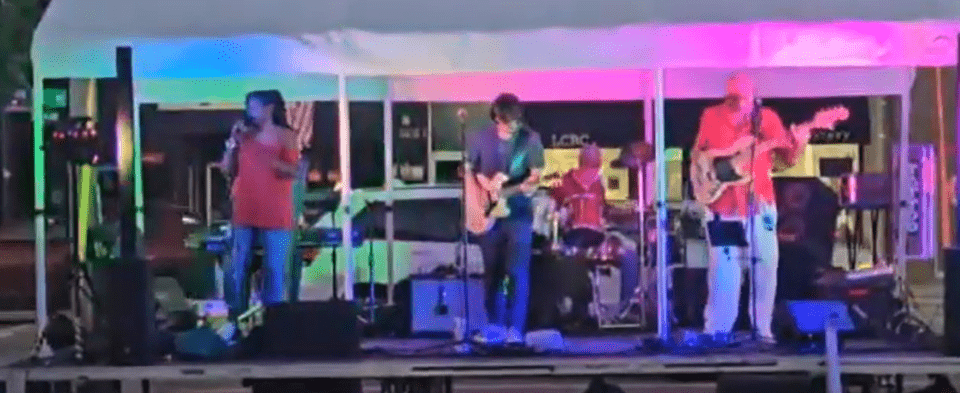 A live band performing under a tent, featuring a singer, guitarist, bassist, and drummer, with colorful stage lighting.
