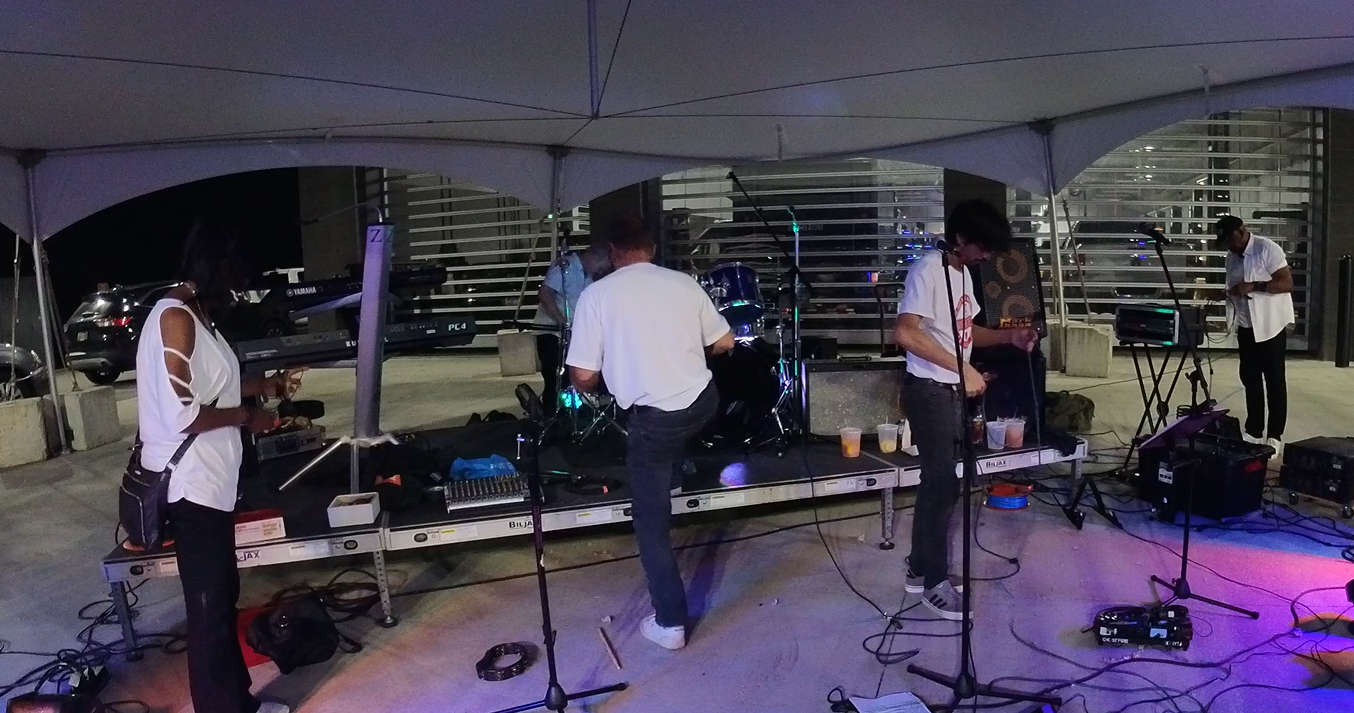 A band performing on a stage under a tent, with musicians playing instruments and a backdrop of parked cars at night.